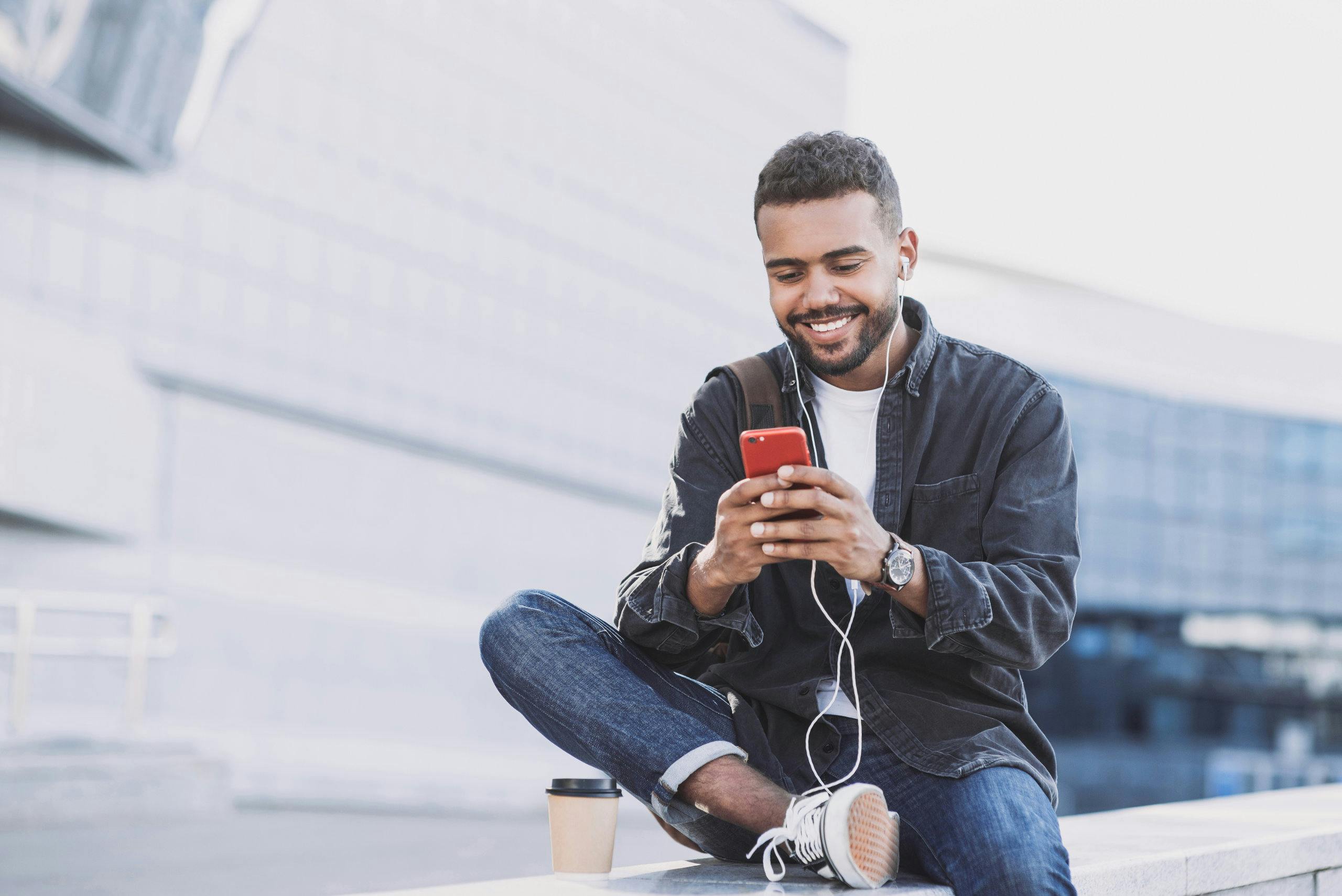 Man holding mobile hone sitting on concrete steps with coffee, smiling at device
